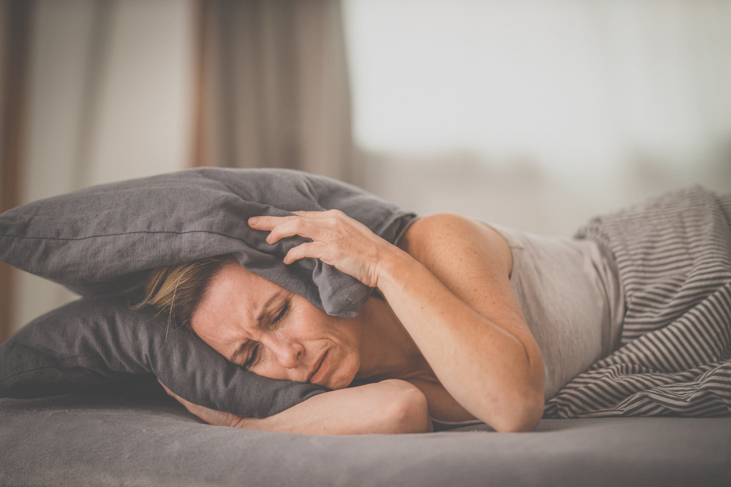 Lady laying in bed covering her ears with pillow, struggling to sleep because of noise.