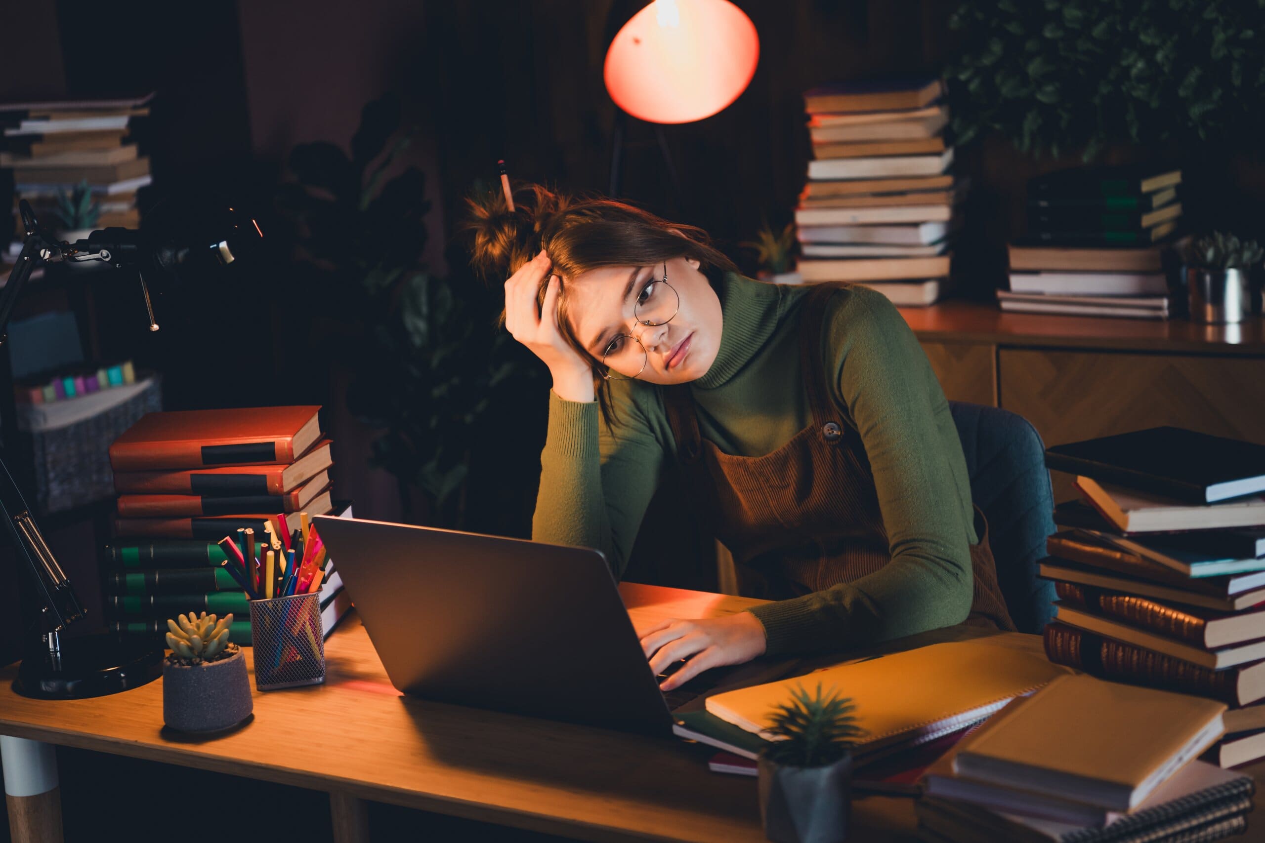 Girl sitting at desk in the dark frowning at laptop, surrounded by books.