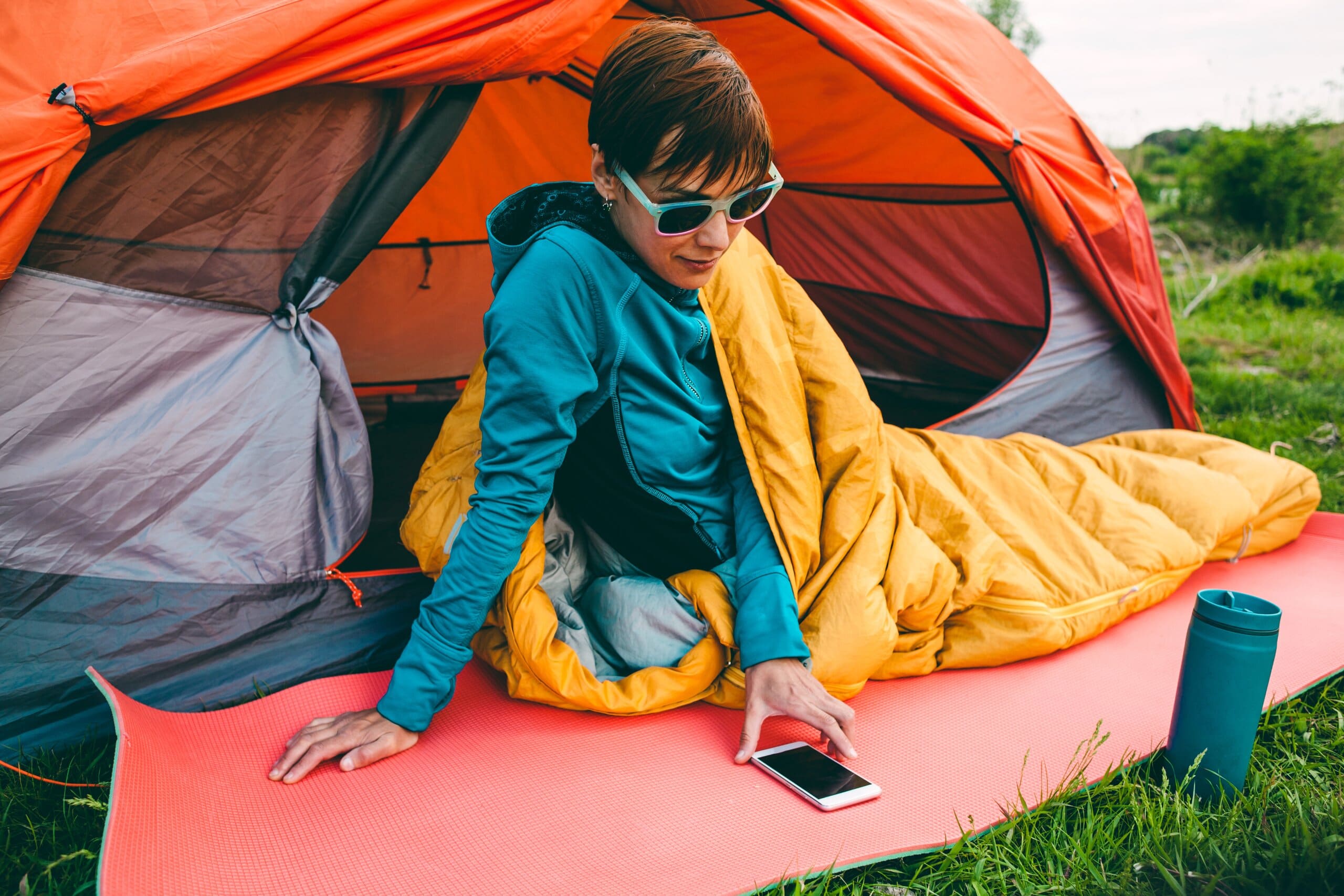 Woman sat up in sleeping bag outside of a tent, checking her phone.