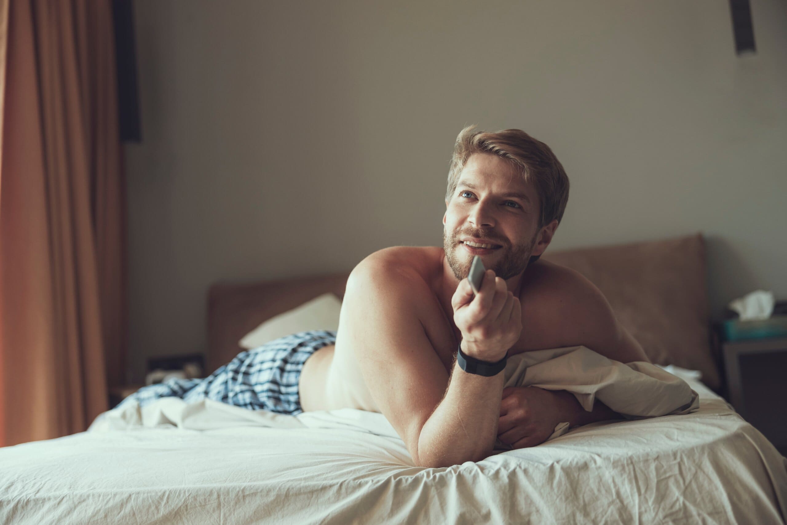 Man lying propped up on his front in bed, holding a TV remote and smiling.