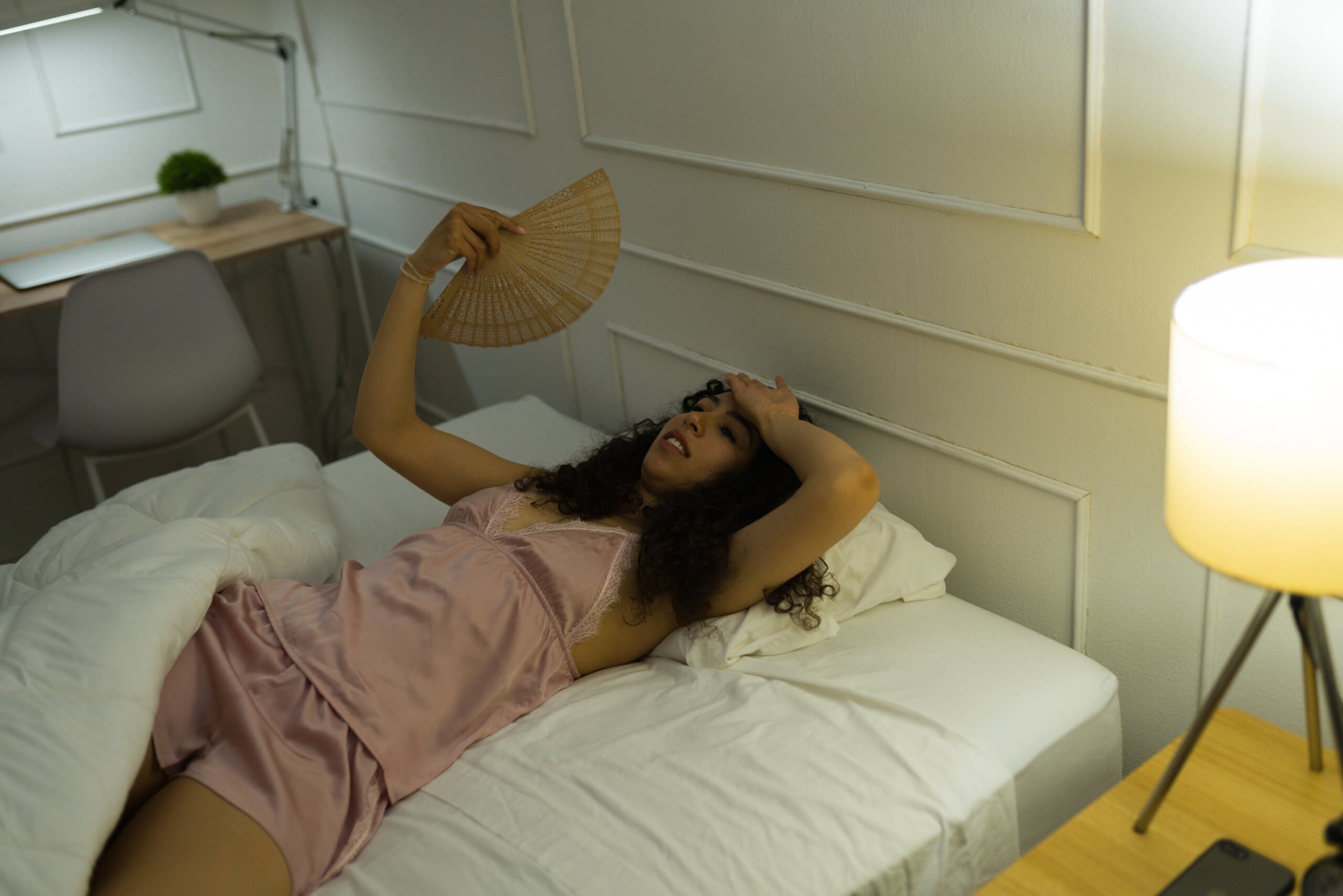 Woman laying in bed struggling to sleep in the heat, fanning herself.