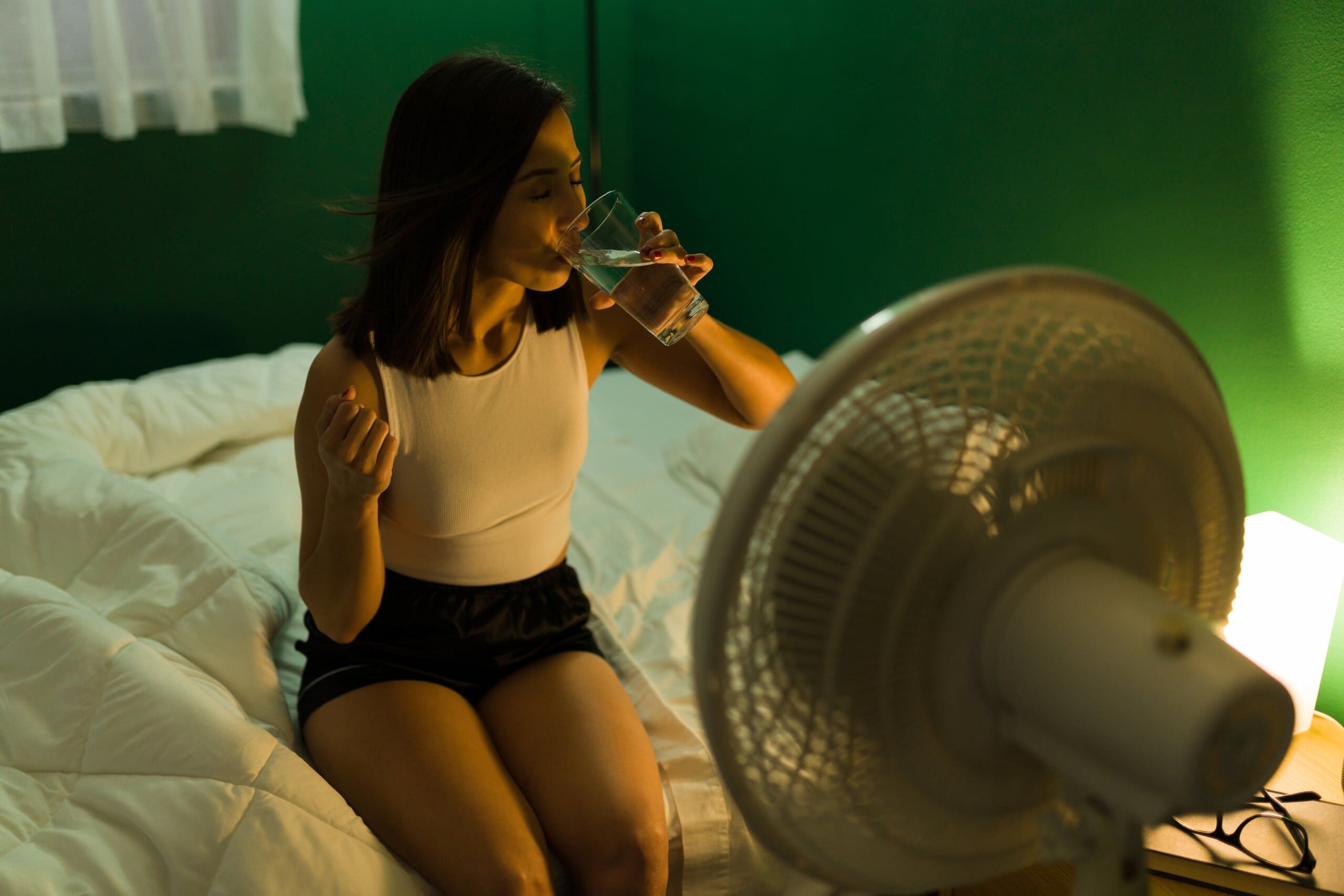 Woman sat on the side of her bed, drinking a glass of water in front of a fan.