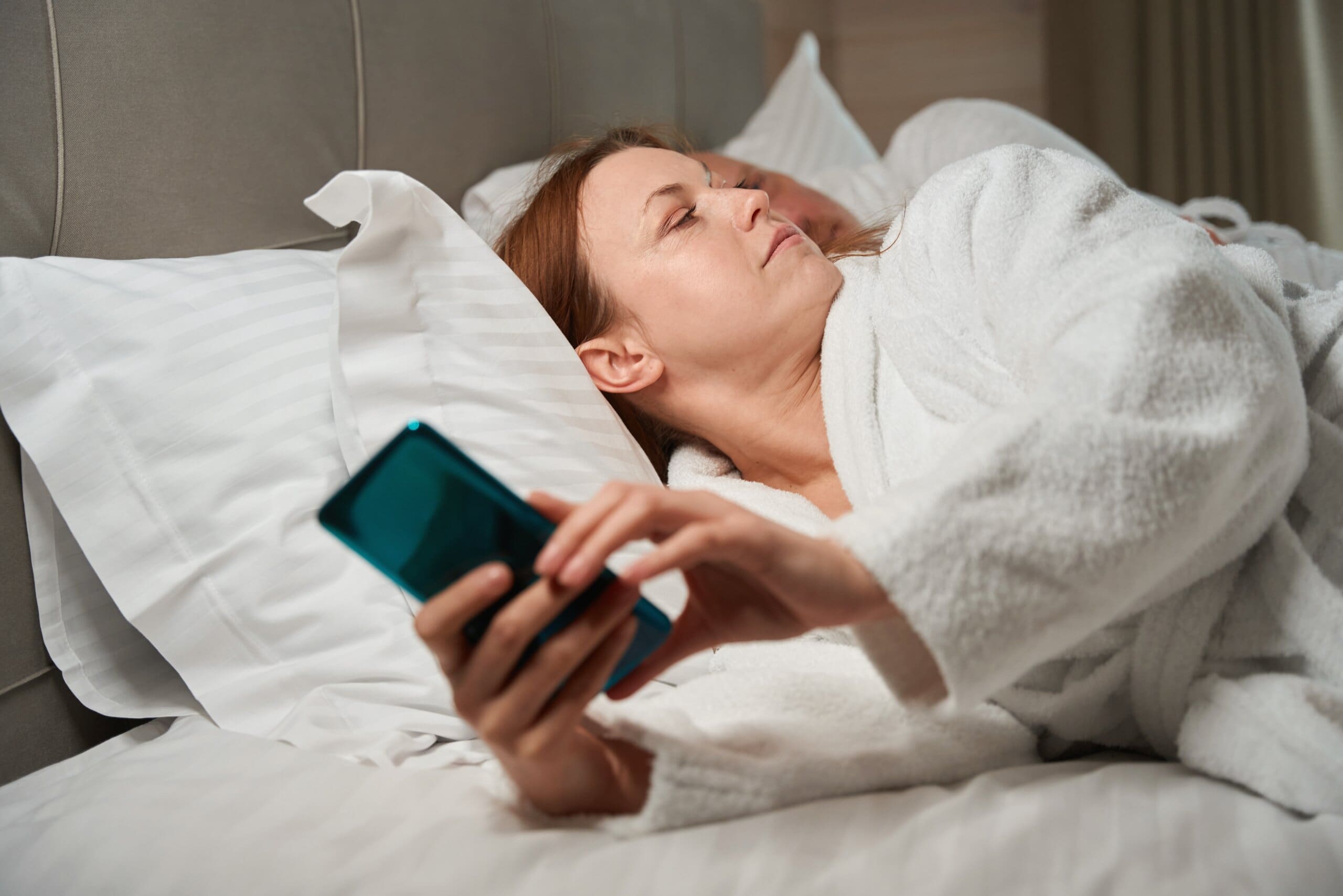 Lady lying in bed on her phone with partner lying next to her.