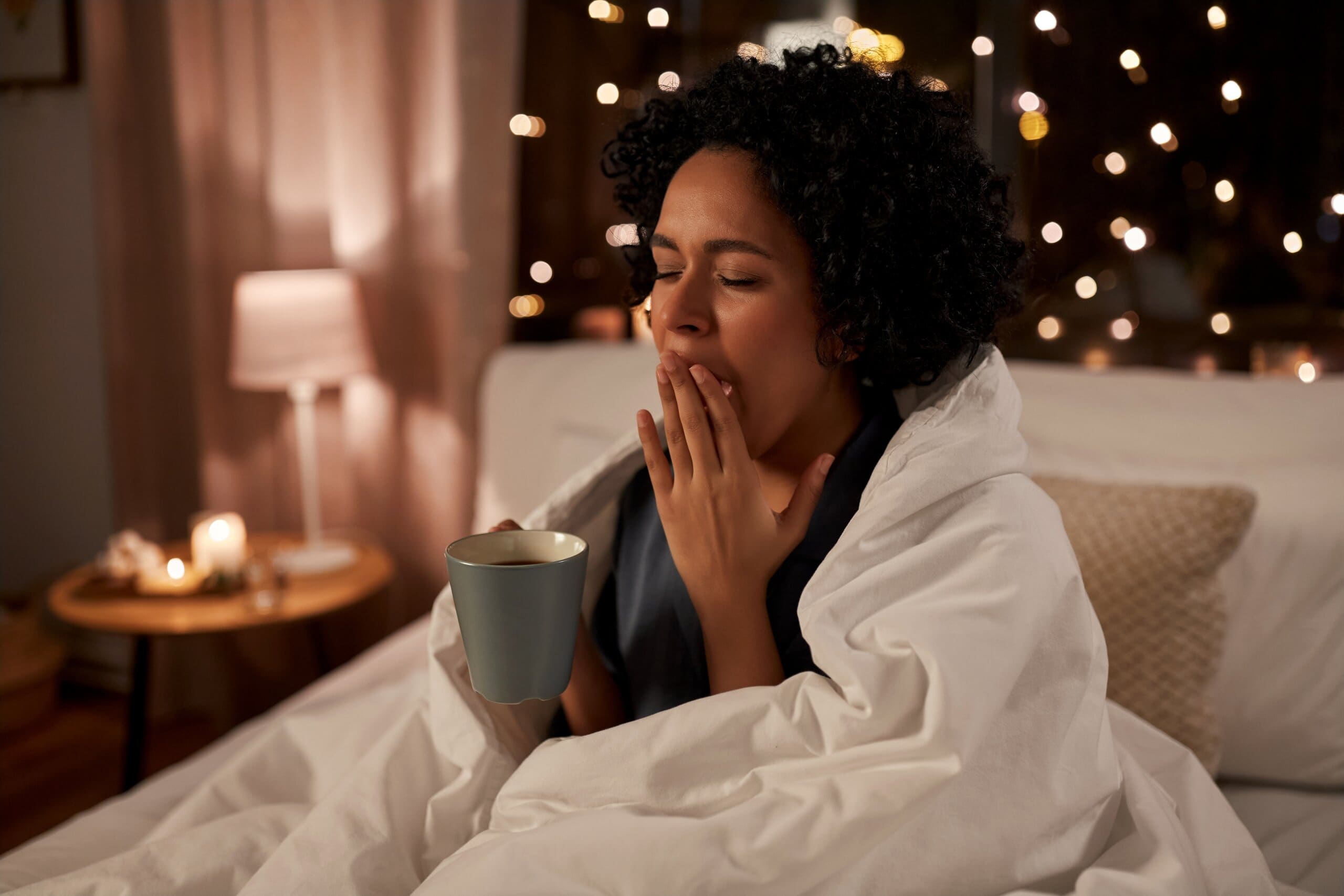 Woman sat up in bed holding a hot drink, yawning.